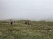 Wanderer in bunten Jacken gehen bei nebligem, bedecktem Wetter einen grasbewachsenen Hügel hinauf.